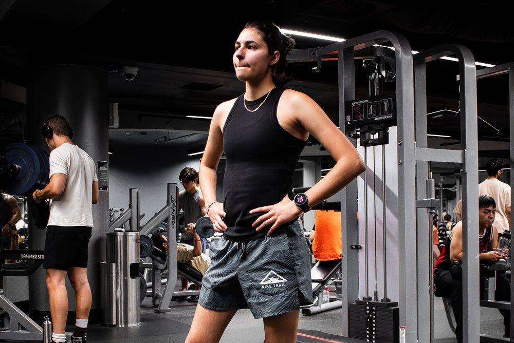 A woman in a gym wearing a black tank top and gray shorts with a white logo, standing in front of a cable machine. Other gym equipment and people are visible in the background. At RMIT Univerity Active Hub