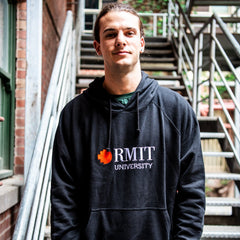 Male model wearing a black Fairtrade hoodie from the RMIT Store, facing forward with both hands relaxed alongside his body. The hoodie features a red and white embroidered RMIT University logo centred on the chest.