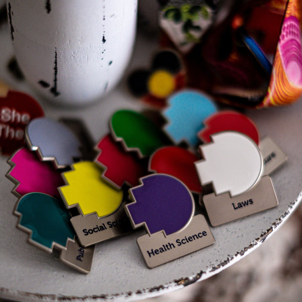 Assorted RMIT Discipline Lapel Matte Pins in multiple colours and fields, arranged overlapping on a distressed white circular stand beside a lanyard and a pronoun pin, with pearls and lace underneath. Available at the RMIT Store.