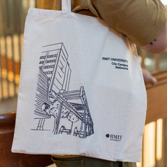 Side view of a model carrying the RMIT Store Everyday Campus Tote in natural cotton, featuring a detailed black line art illustration of the RMIT City Campus. The artwork includes the iconic Swanston Academic Building with its angular architecture and surrounding street-level details. The RMIT University logo and “City Campus” are printed above the illustration in black. The model wears a tan polo shirt and olive green cargo pants, photographed indoors with warm lighting.