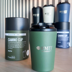 Everyday Reusable Cup in Khaki with a matte green finish, black screw-on sip lid and silver RMIT University logo on the front, displayed beside its cylindrical Fressko Café Collection Khaki packaging on a timber table, with striped wall panelling and a warm glass wall light in the background at RMIT Store.