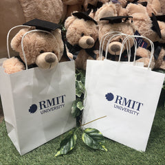 Two white RMIT University gift bags with twisted handles standing on artificial grass in front of a display of graduation teddy bears wearing black mortarboards inside the RMIT Store.