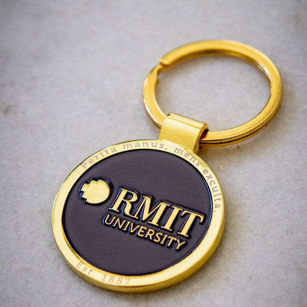 Angled front view of the RMIT motto lapel keyring, featuring a navy centre with a gold outer edge and gold RMIT branding, attached to a gold circular handle, displayed on a light background, available at the RMIT Store.