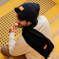 Male model looking back towards the camera while wearing the navy RMIT knitted beanie with a brown PU patch embossed with the RMIT logo, and the matching navy knitted scarf. The model is seated on outdoor stairs in an urban setting, dressed casually in a white crew neck sweater. The knit texture and details of both the beanie and scarf are clearly visible. Available at the RMIT Store.