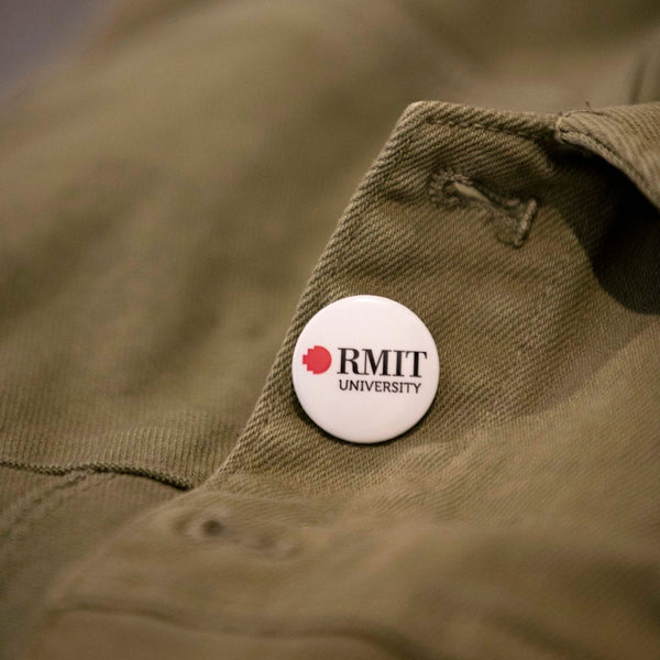 A white RMIT University badge on a green jacket. The badge displays the RMIT logo in white and red, prominently positioned on the left chest area of the jacket.