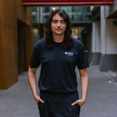 Model standing in an outdoor campus walkway wearing RMIT Essential Tee in navy featuring a small white RMIT University logo printed on the left chest, relaxed classic fit with short sleeves, paired with black pants and hands in pockets in a modern architectural setting, available at the RMIT Store.