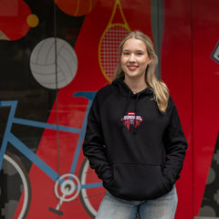 Female model wearing a black Redbacks Hoodie with embroidered red and white Redbacks spider logo on the chest, front kangaroo pocket and black drawstrings, standing in front of a colourful sports-themed wall featuring basketball, tennis and cycling graphics, available at the RMIT Store.