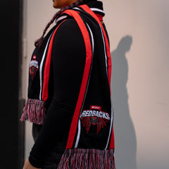 Model wearing the Redbacks scarf with bold red, black, and white stripes, featuring an embroidered Redbacks logo and spider design along with the RMIT branding. The scarf has multicoloured fringes at the ends, and is worn over a black top in a side studio setting. Highlighting the scarf's distinct RMIT Store branding detail.