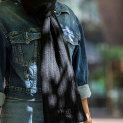 A model wearing the RMIT Ribbed Scarf, styled with a denim jacket and jeans. The black ribbed scarf is elegantly wrapped around the neck, its texture and warmth highlighted by the soft sunlight filtering through leaves.