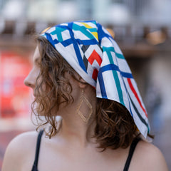 Female model wearing the Sentient Scarf styled as a headscarf, featuring abstract geometric artwork in blue, teal, red, yellow, black and white with satin finish fabric, paired with gold geometric earrings and a black ribbed camisole in an outdoor urban setting, available at the RMIT Store.