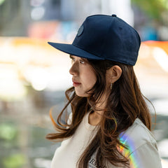 Side view of RMIT Store snapback cap in navy blue with embroidered logo, worn by a model with long, wavy hair and gold hoop earrings. The cap complements the casual outfit in an outdoor urban setting.
