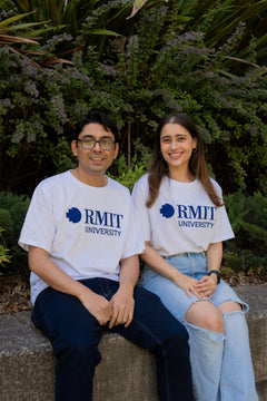 Front view of two models sitting on a stone bench wearing plain white RMIT Welcome tees from the RMIT Store, each featuring a bold blue RMIT logo printed centrally on the chest. The male model on the left wears dark denim jeans, while the female model on the right wears light blue denim jeans. Both sit casually against a neutral outdoor background, highlighting the relaxed fit and clean design of the tees.