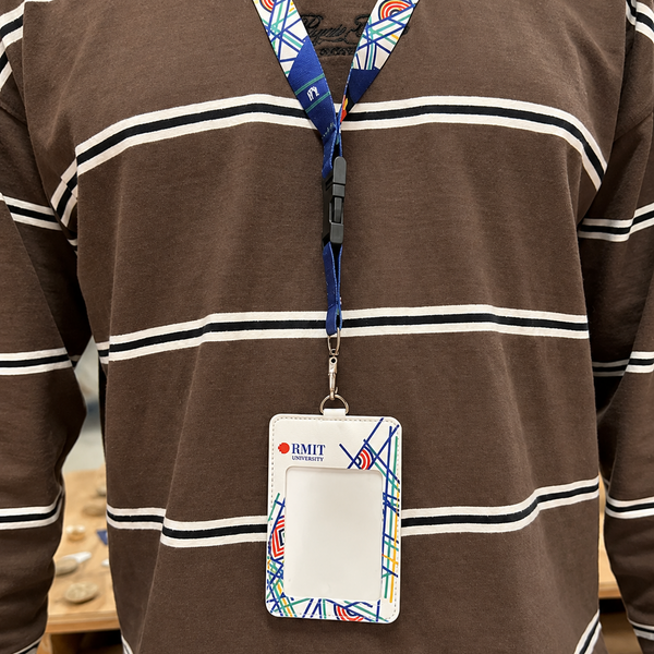 White Sentient Card Holder front view worn on a patterned RMIT lanyard by a model in a brown striped long sleeve top, showing colourful geometric artwork and small printed flags, available at the RMIT Store.