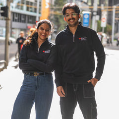 Two students wearing RMIT Essential half zip jumper in navy with embroidered RMIT University logo, standing together outdoors in the city and smiling.