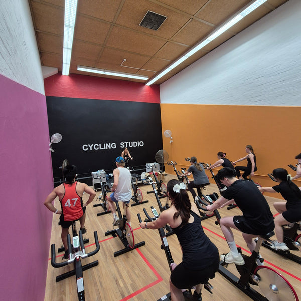 A group of people riding indoor stationary bikes in a cycling studio at the RMIT Sports Centre with wood floors, a black back wall with white text, and pink, red, and orange painted walls.