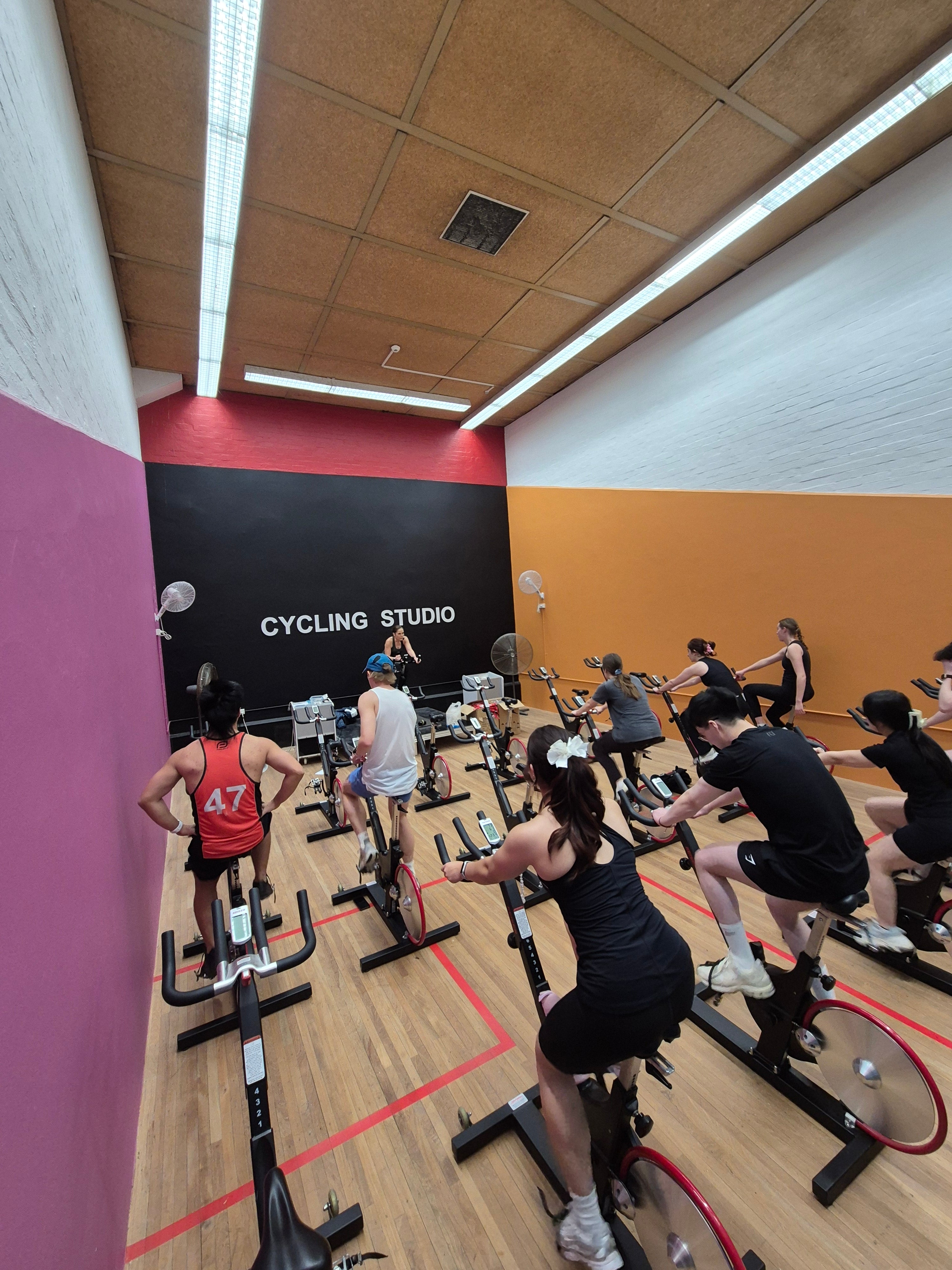 A group of people riding indoor stationary bikes in a cycling studio at the RMIT Sports Centre with wood floors, a black back wall with white text, and pink, red, and orange painted walls.