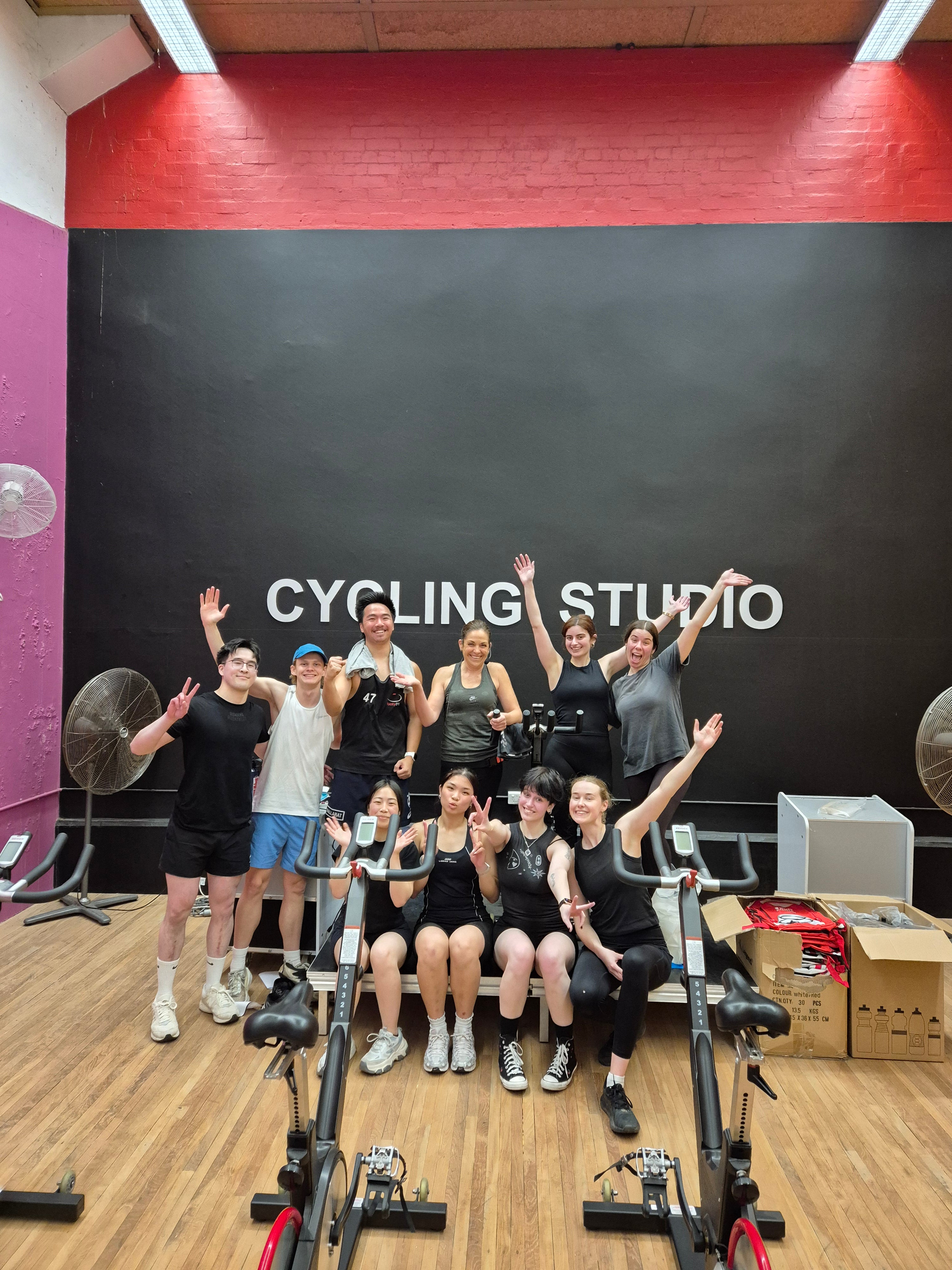 A group of nine fitness enthusiasts posing together in a gym with a large black wall that reads 'CYCLING STUDIO'. They are gathered around and in front of several stationary exercise bikes at the RMIT Sports Centre.