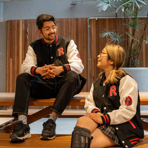 Two people wearing matching RMIT University varsity jackets sitting on a bench indoors.