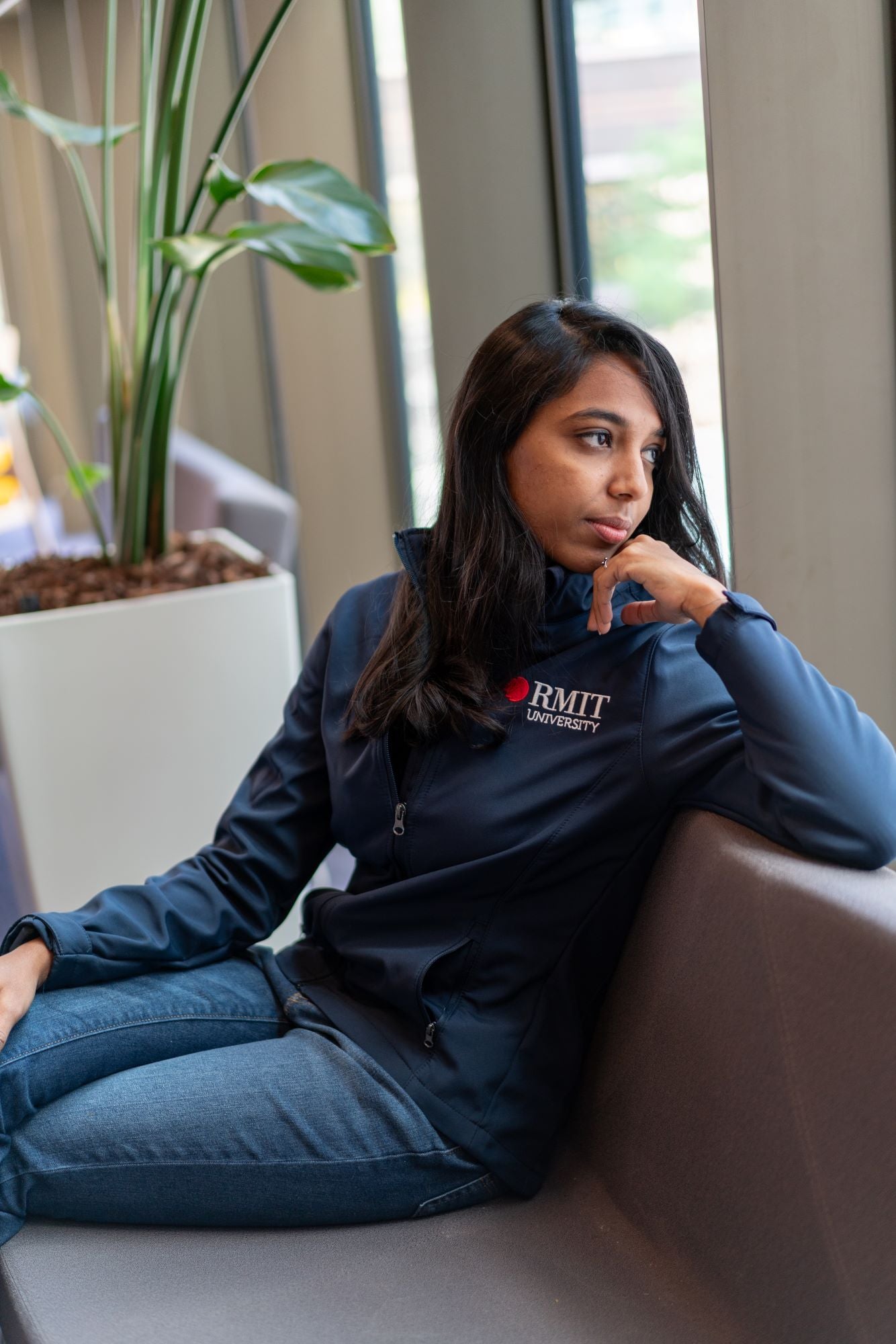 Female model seated casually in an urban environment, wearing the RMIT Softshell Jacket in navy. The jacket is half unzipped, revealing a black top underneath. The model’s left hand is resting under her chin, and her legs are relaxed. The embroidered RMIT logo is visible on the left chest of the jacket. The scene highlights the jacket’s structured fit, high collar, and everyday wear styling. Available at the RMIT Store.