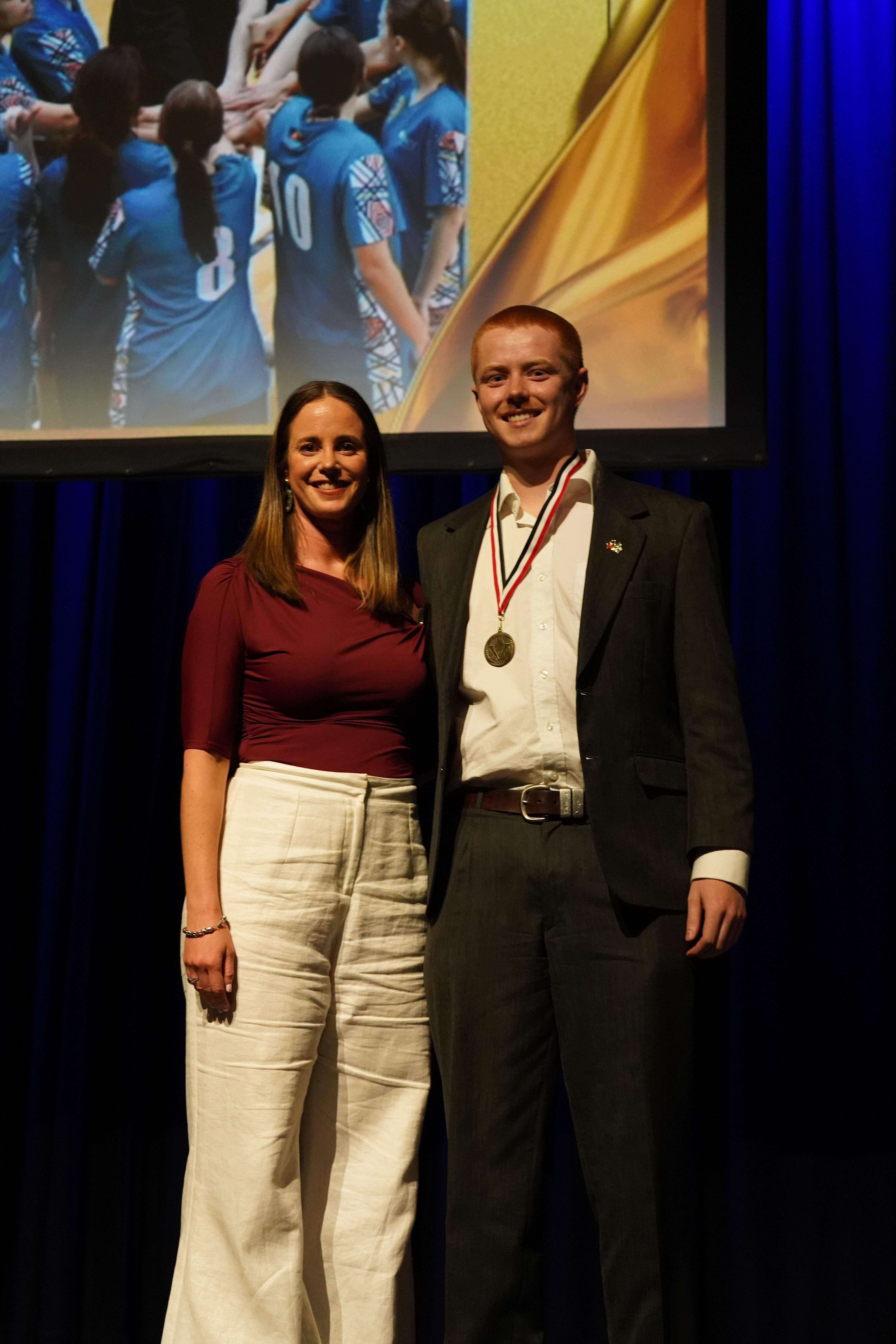 A man and woman posing on stage at the RMIT Active Awards Night, with a screen behind them showing a sports team photo