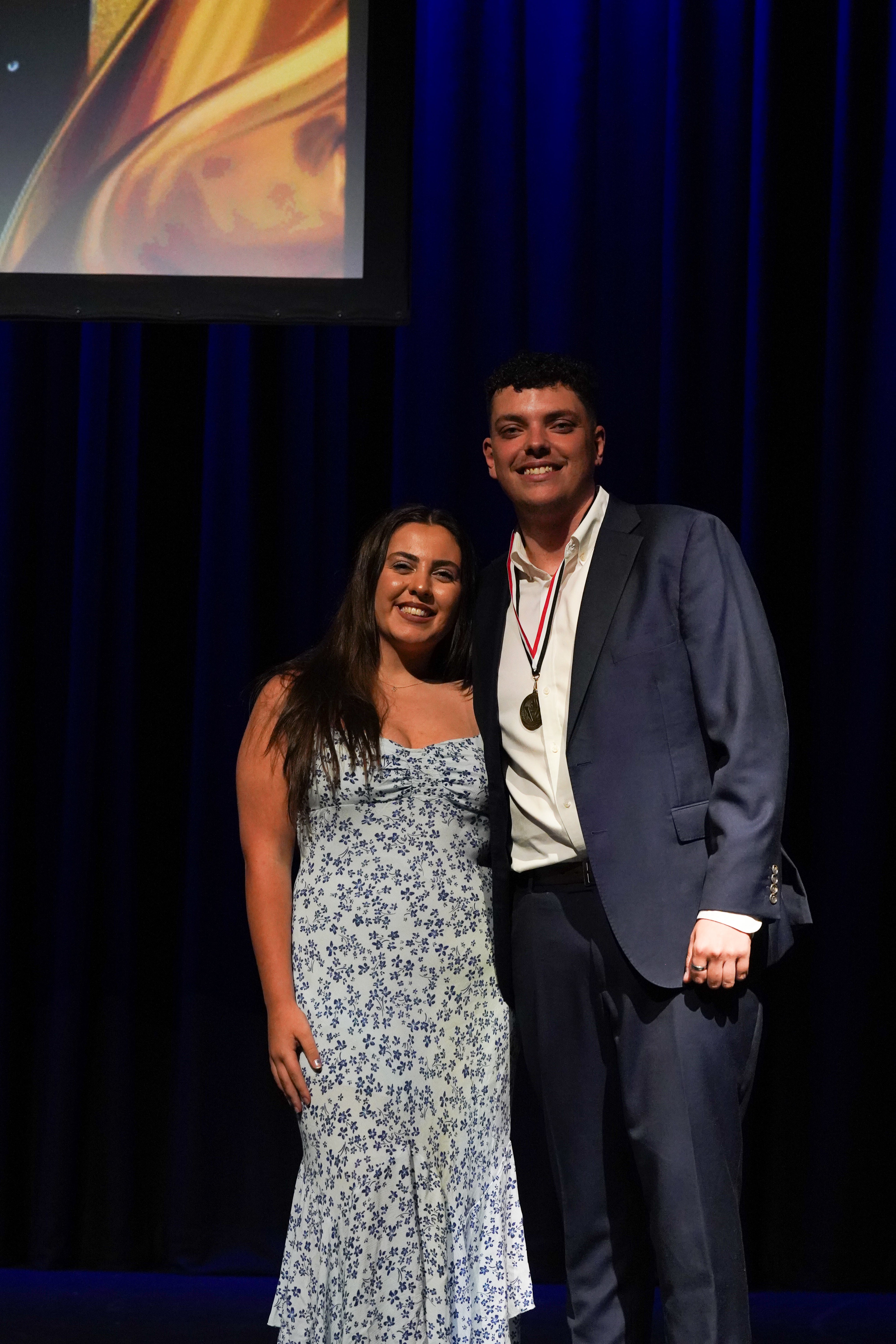 A man wearing a medal stands on stage with a woman in a floral dress at the RMIT Active Awards Night