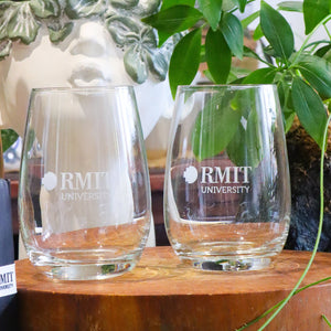 Two clear glass tumblers with the RMIT University logo frosted on the surface, placed on a wooden surface with foliage in the background.