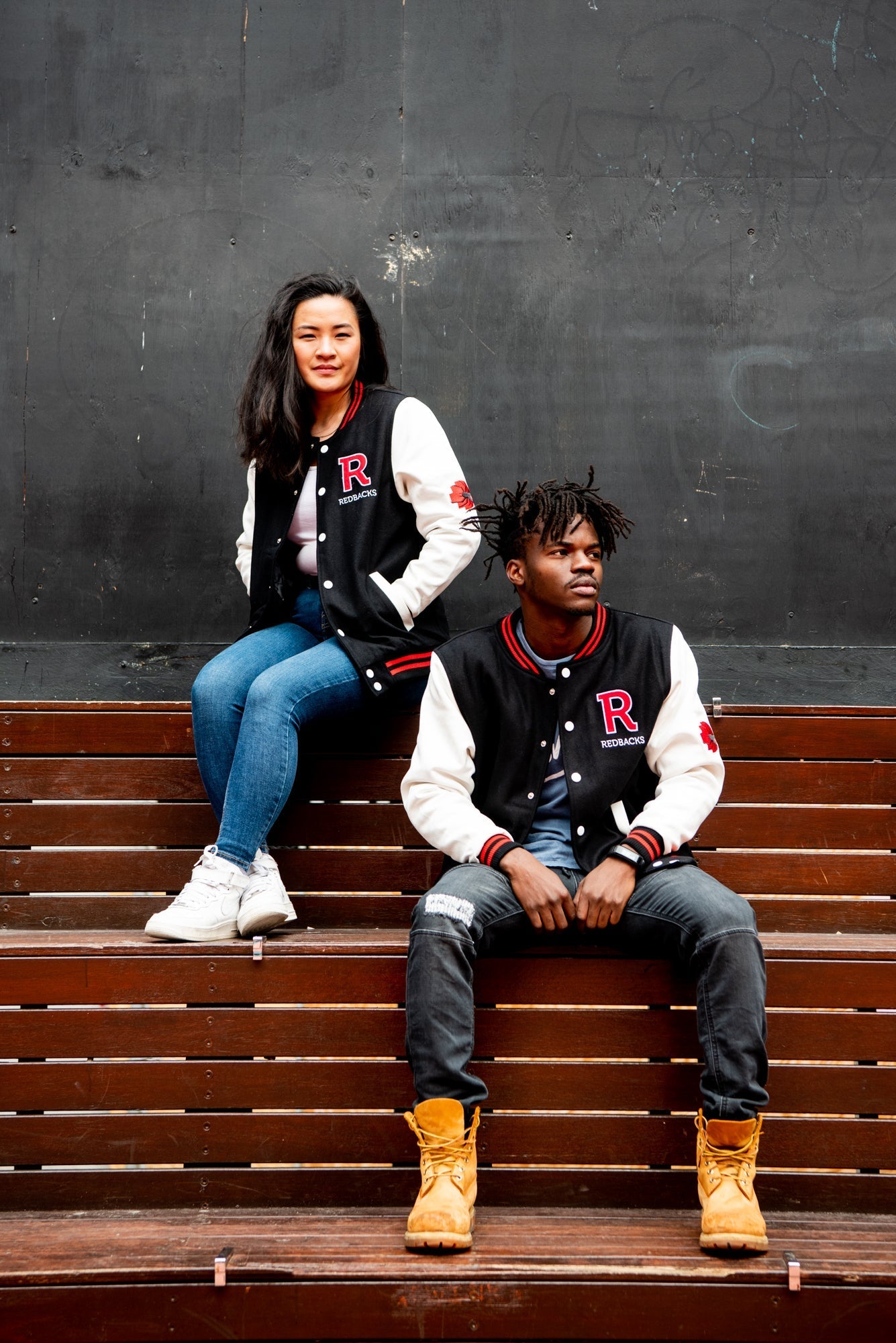 Male and female models wear unbuttoned RMIT Store black varsity jackets with white sleeves, black collars and hems with red stripe detailing, and large red chenille letter "R" patches on the left chest; seated side by side on a wooden bench in an outdoor on-campus setting, the female model is on the left wearing light blue denim jeans, and the male model is on the right wearing dark denim jeans.