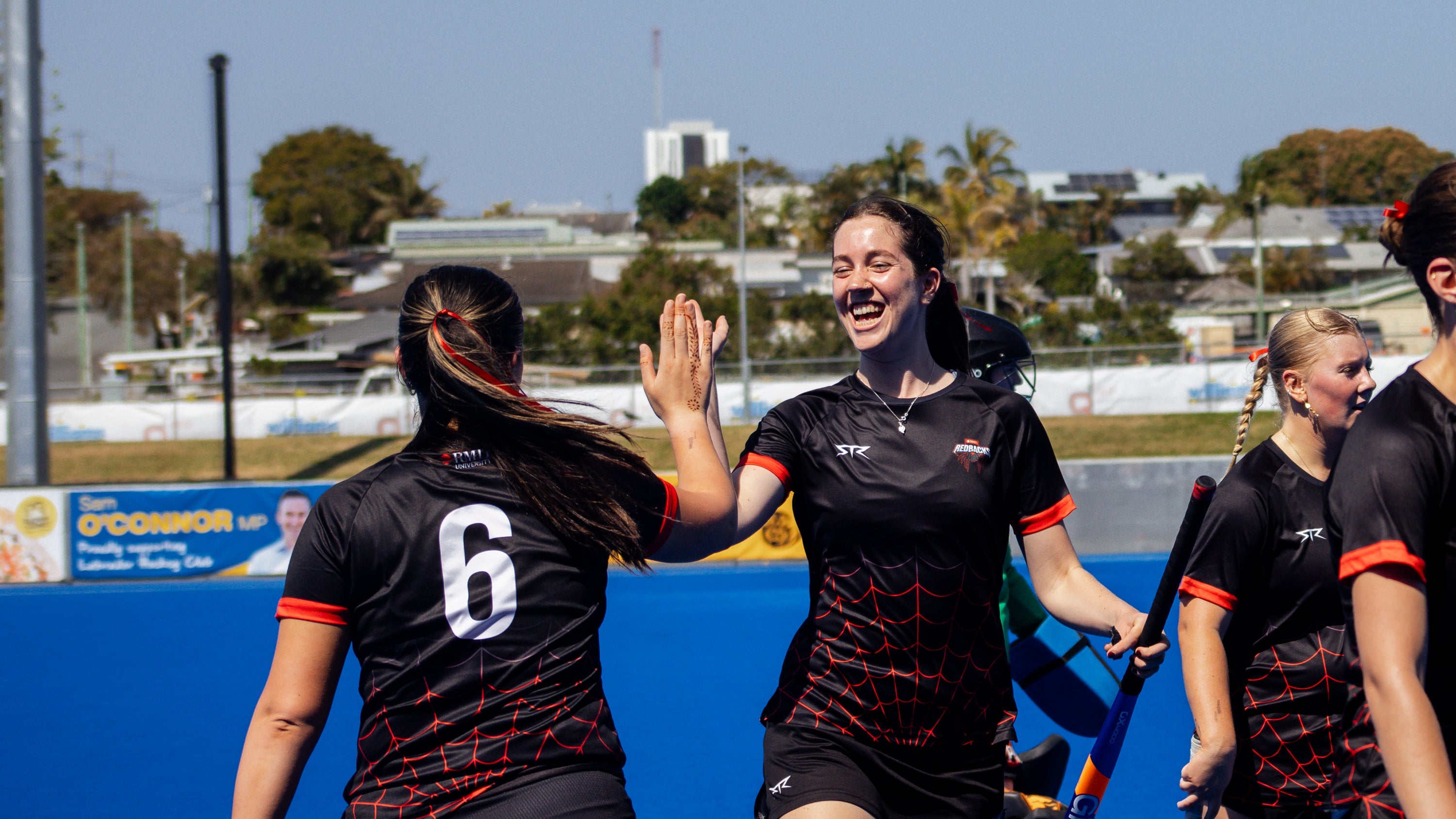 Two female athletes in black RMIT University Redbackssports uniforms high-fiving on a blue field with stadium in the background