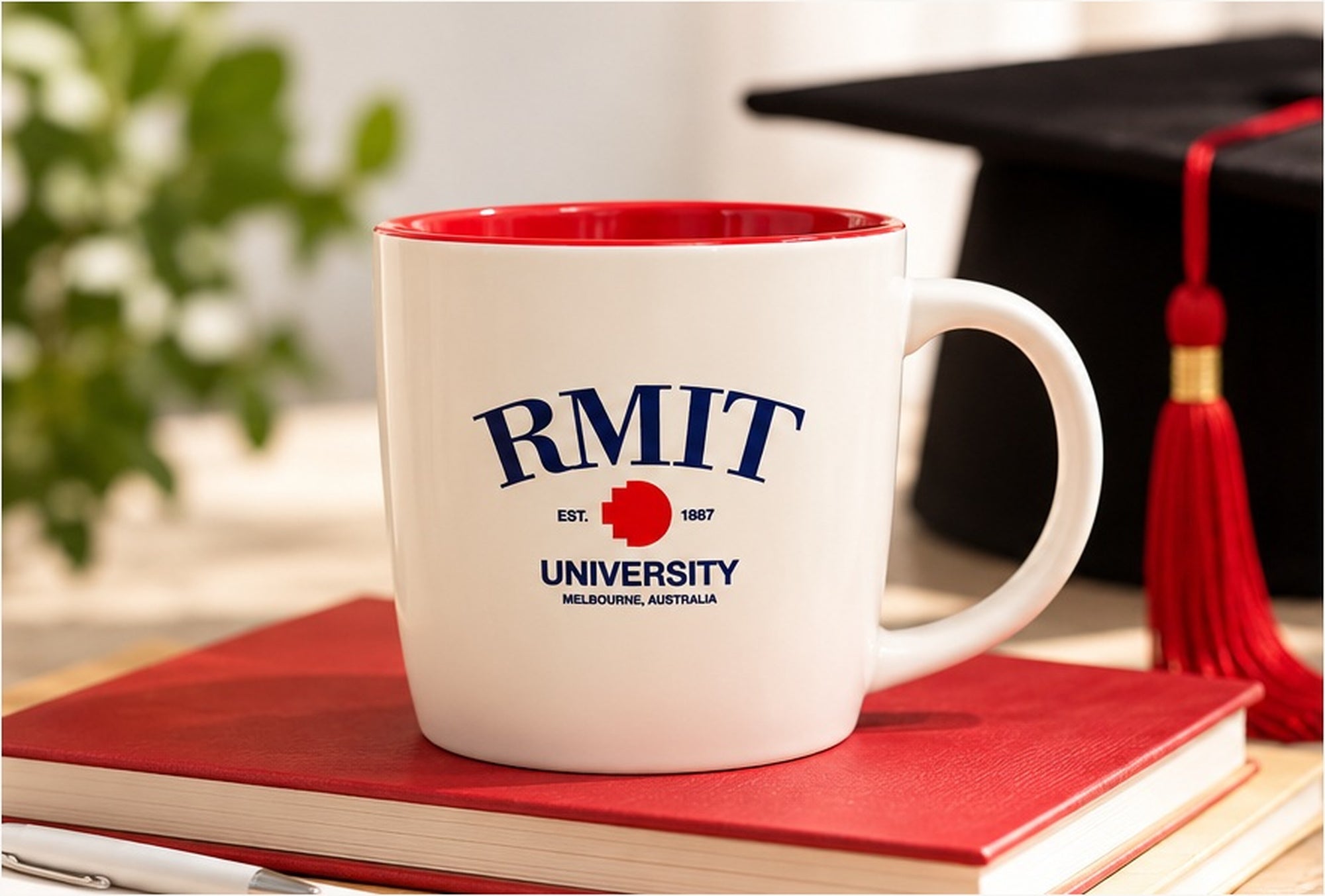 RMIT 1887 Mug in white ceramic with a glossy red interior, featuring navy RMIT University lettering, a red pixel logo and EST. 1887 detailing. Displayed on a red hardcover book beside a black graduation cap with a red tassel, against a softly blurred background for RMIT Store.