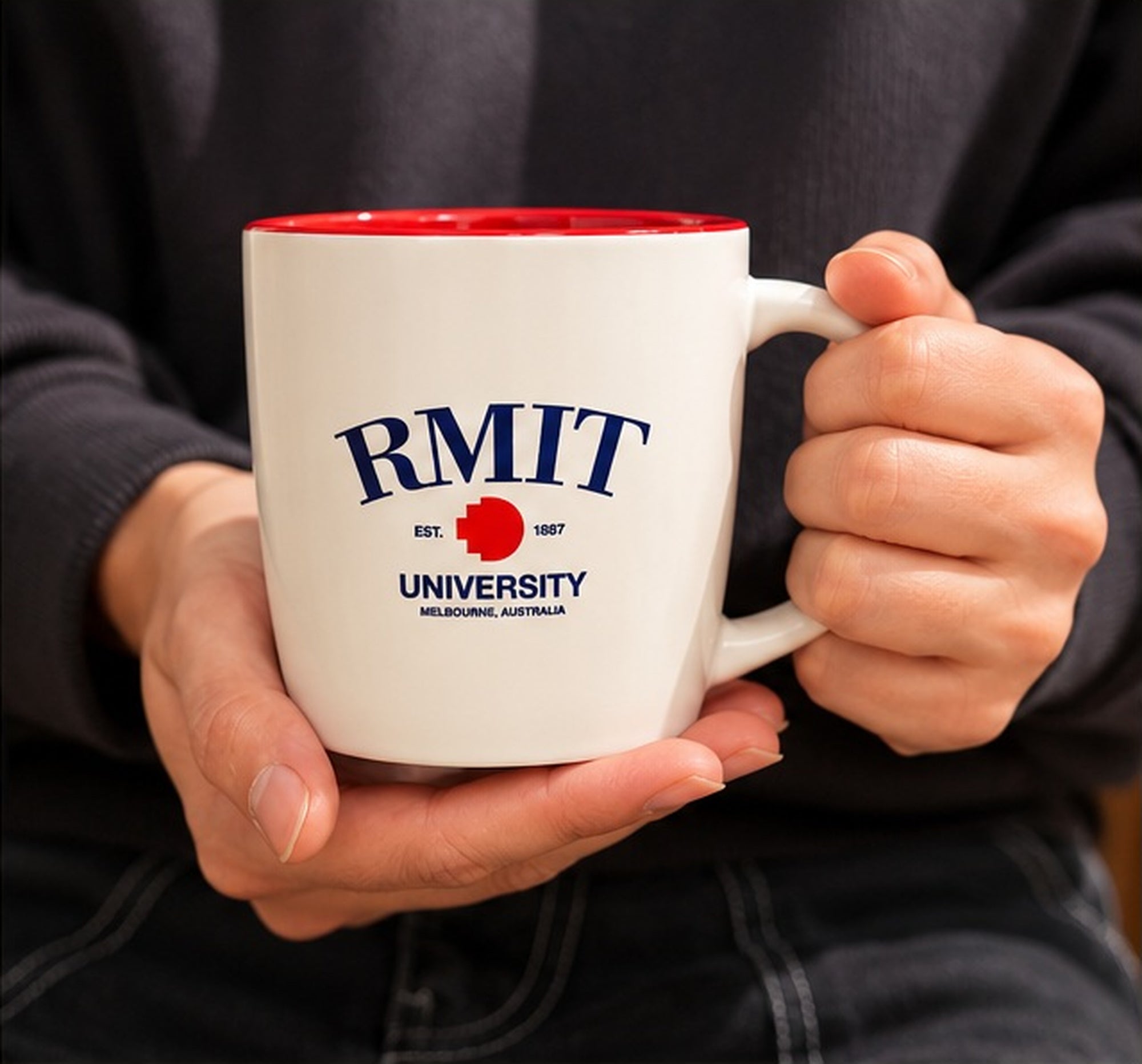 RMIT 1887 Mug in white ceramic with a red interior, held by a seated model wearing a dark sweatshirt and trousers. The model grips the white handle with one hand and supports the base with the other, displaying navy RMIT University branding, a red pixel logo and EST. 1887 detailing for RMIT Store.