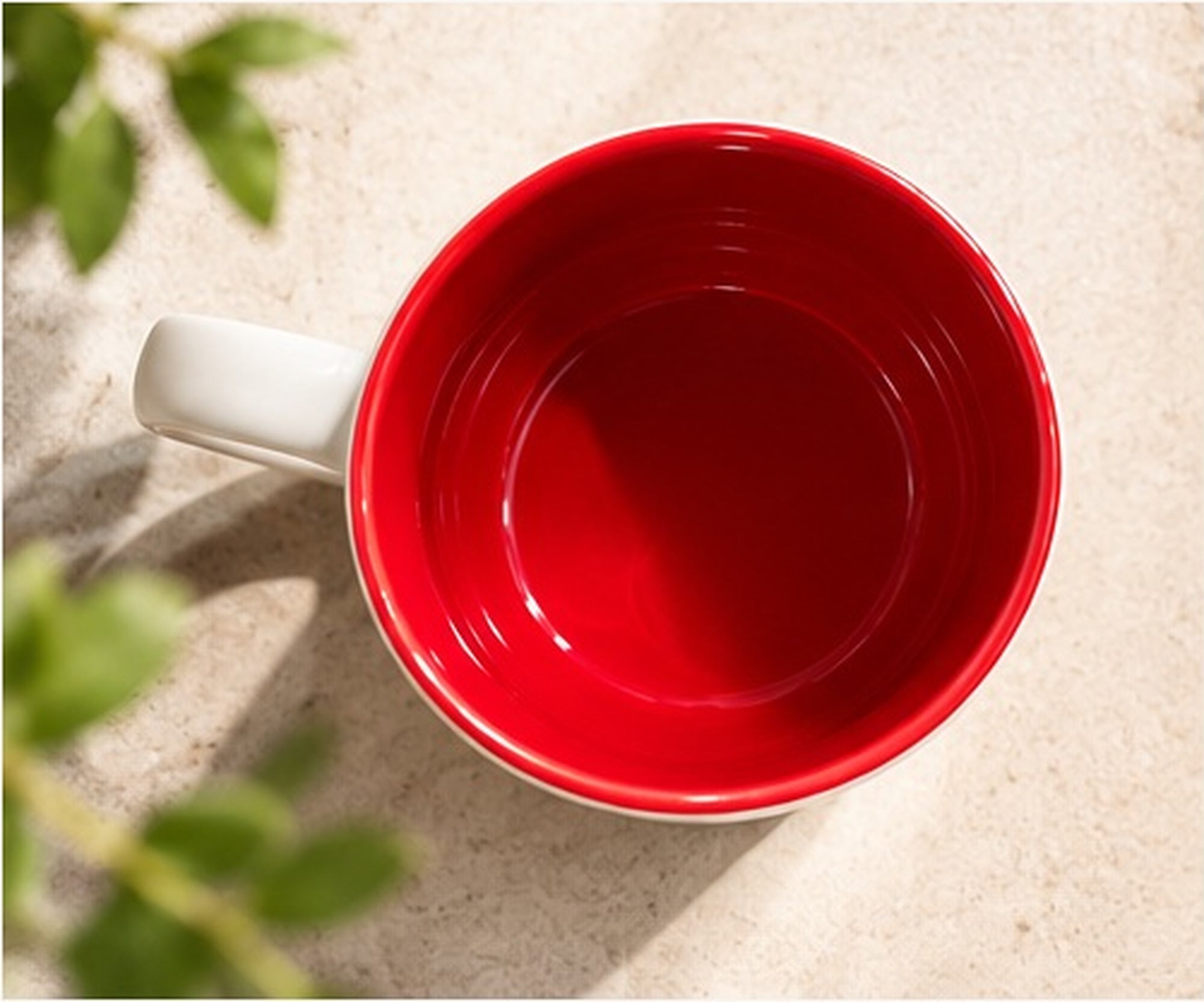 Overhead view of the RMIT 1887 Mug, showing its glossy deep-red interior, matching red rim and white ceramic handle extending to the left. Photographed on a light stone surface with soft natural shadows and blurred green foliage for RMIT Store. Exterior branding is not visible.