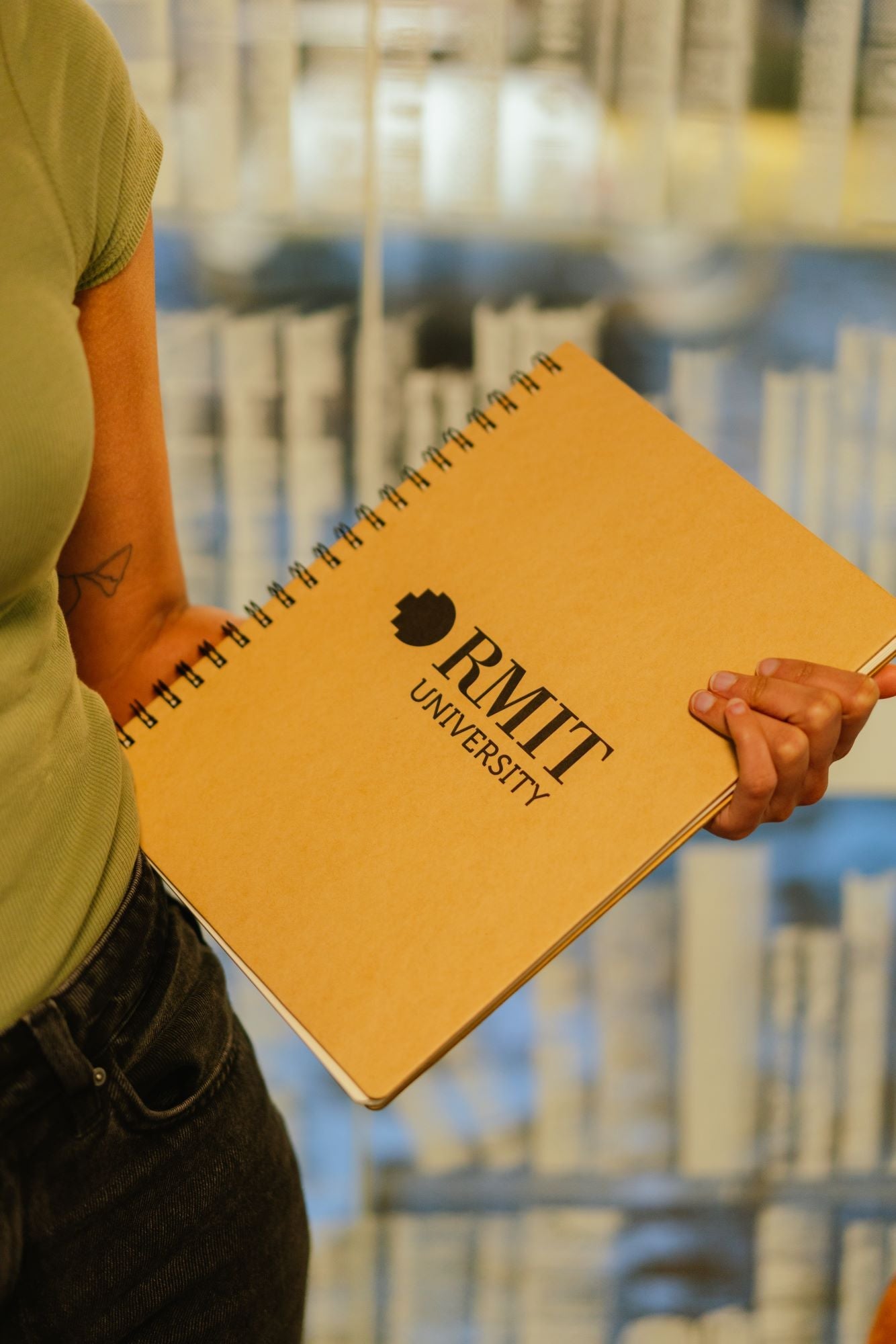 Close-up of a model holding an A4 recycled notebook from the RMIT Store with both hands, displaying the front cover. The notebook features a black RMIT logo printed in the centre of its brown kraft paper cover, with a visible natural recycled texture.