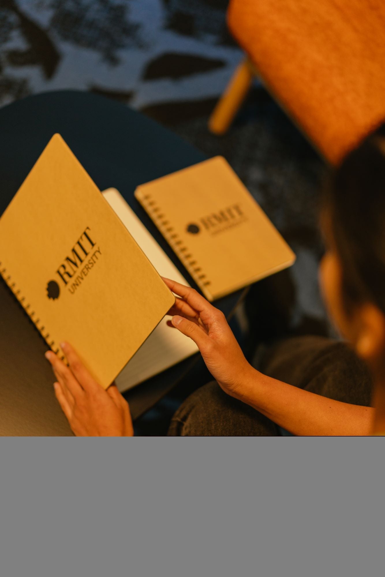 A model seated at a table opening an A4 recycled notebook from the RMIT Store, revealing lined pages. An A5 recycled notebook with a visible black RMIT logo on the cover lies closed next to it. Both notebooks have brown kraft paper covers and rest on a wooden tabletop.