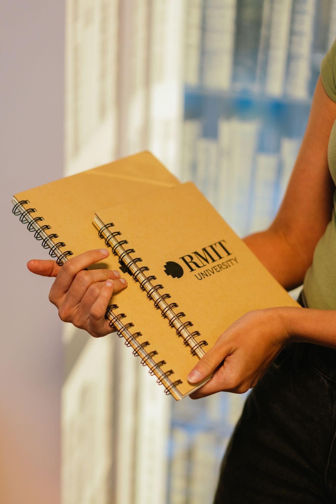 A model’s hands holding a stack of RMIT Store recycled notebooks, with an A5 notebook on top of an A4 notebook. The A5 notebook features a black RMIT logo printed in the centre of its brown kraft paper cover. Both notebooks have a natural recycled texture