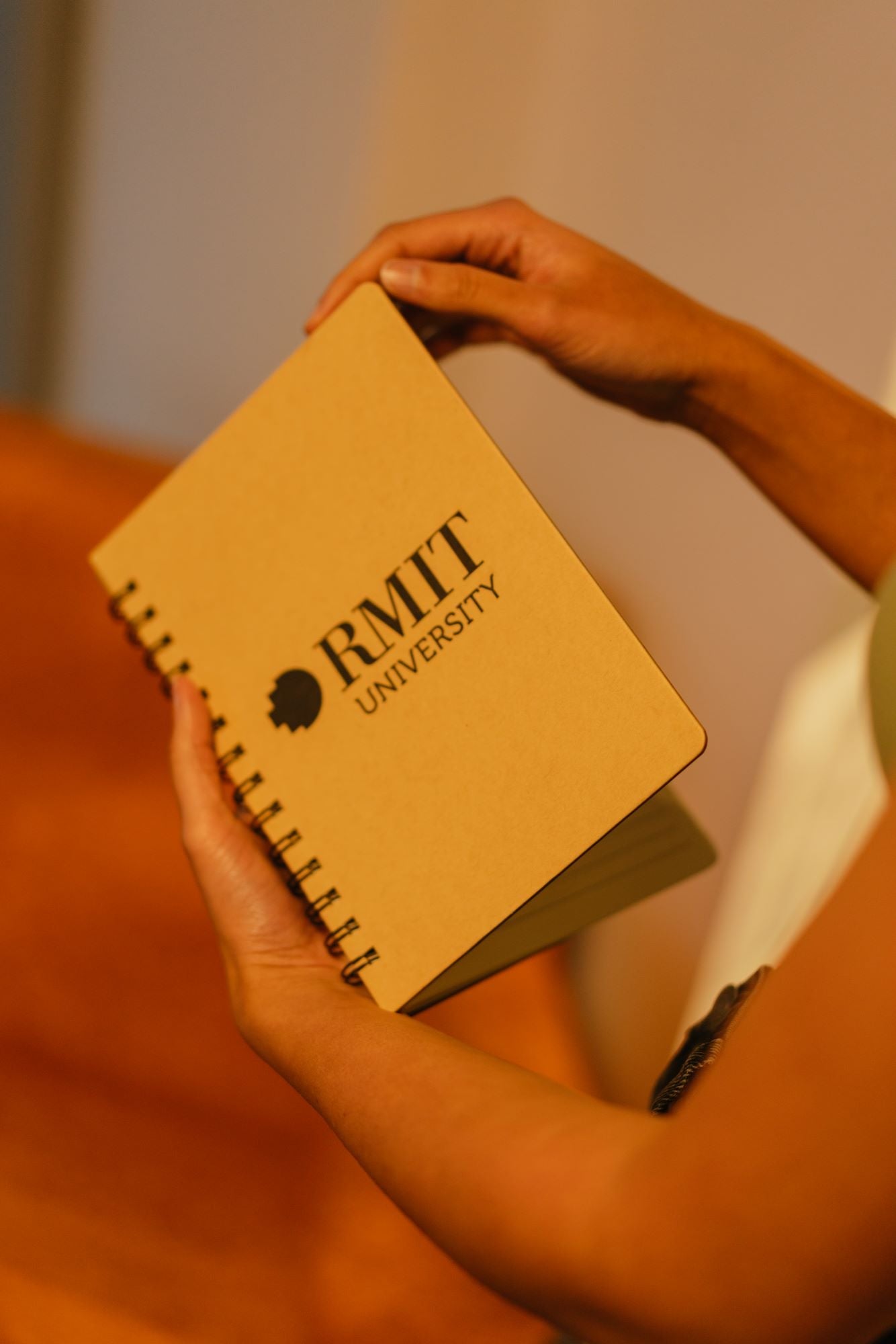 Modelled view of the RMIT Store A5 Recycled Notebook on a wooden tabletop, with the model lifting the brown kraft cover to reveal lined pages. The RMIT University logo is printed in black at the centre of the front cover.