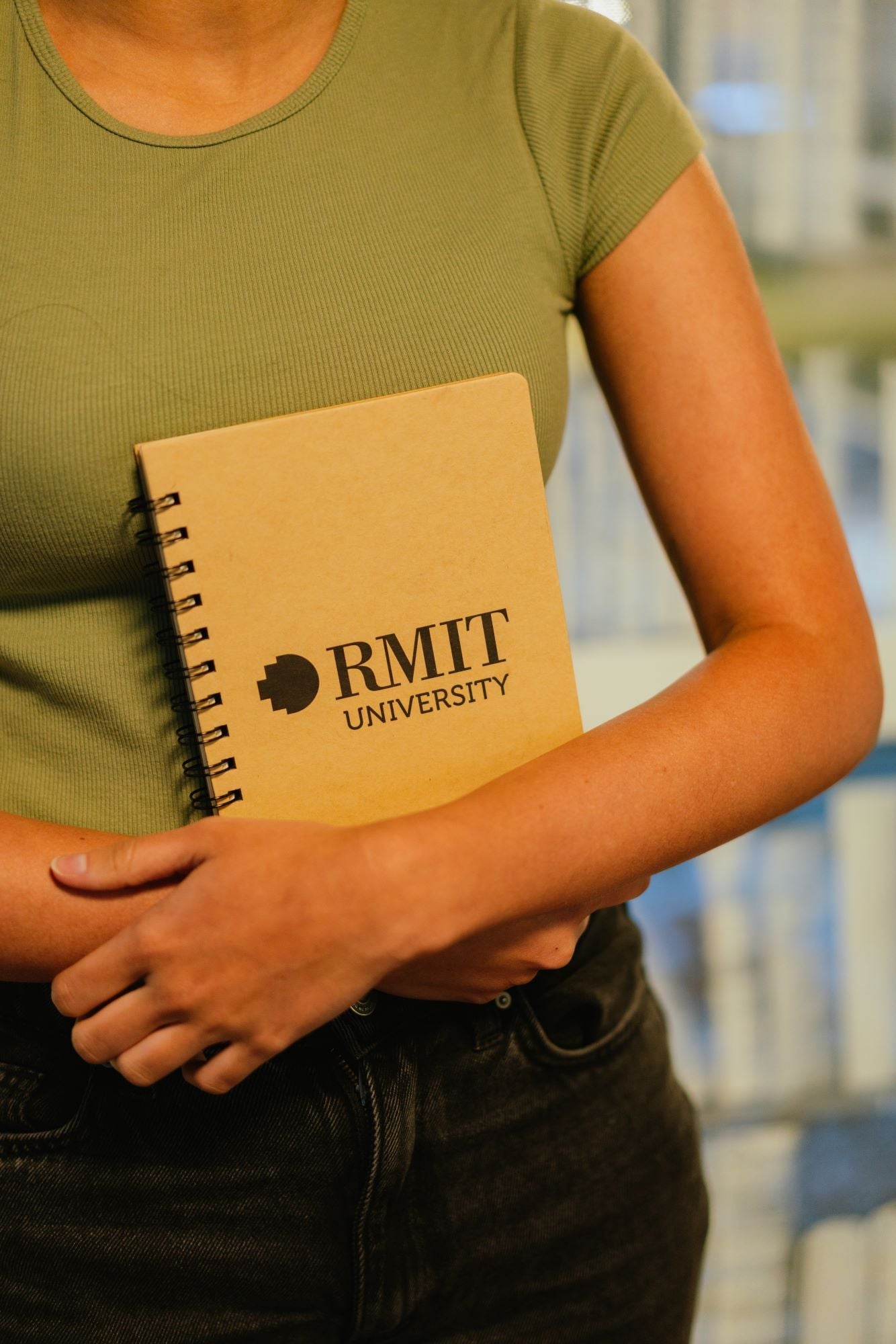 Model holding the closed RMIT Store A5 Recycled Notebook with a brown kraft cover and black spiral binding. The RMIT University logo is clearly printed in black at the centre of the front cover
