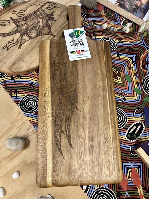 RMIT Store Acacia Rectangle Paddle Board, displayed on a wooden table in-store. The board features a smooth acacia wood surface with natural grain patterns and a rectangular shape with rounded edges. A detailed gum leave design is meticulously etched into the board, inspired by Traditional South Eastern Aboriginal art, adding a cultural touch to this functional and artistic piece. The piece is part of the Ngarga Warendj – Dancing Wombat collection.