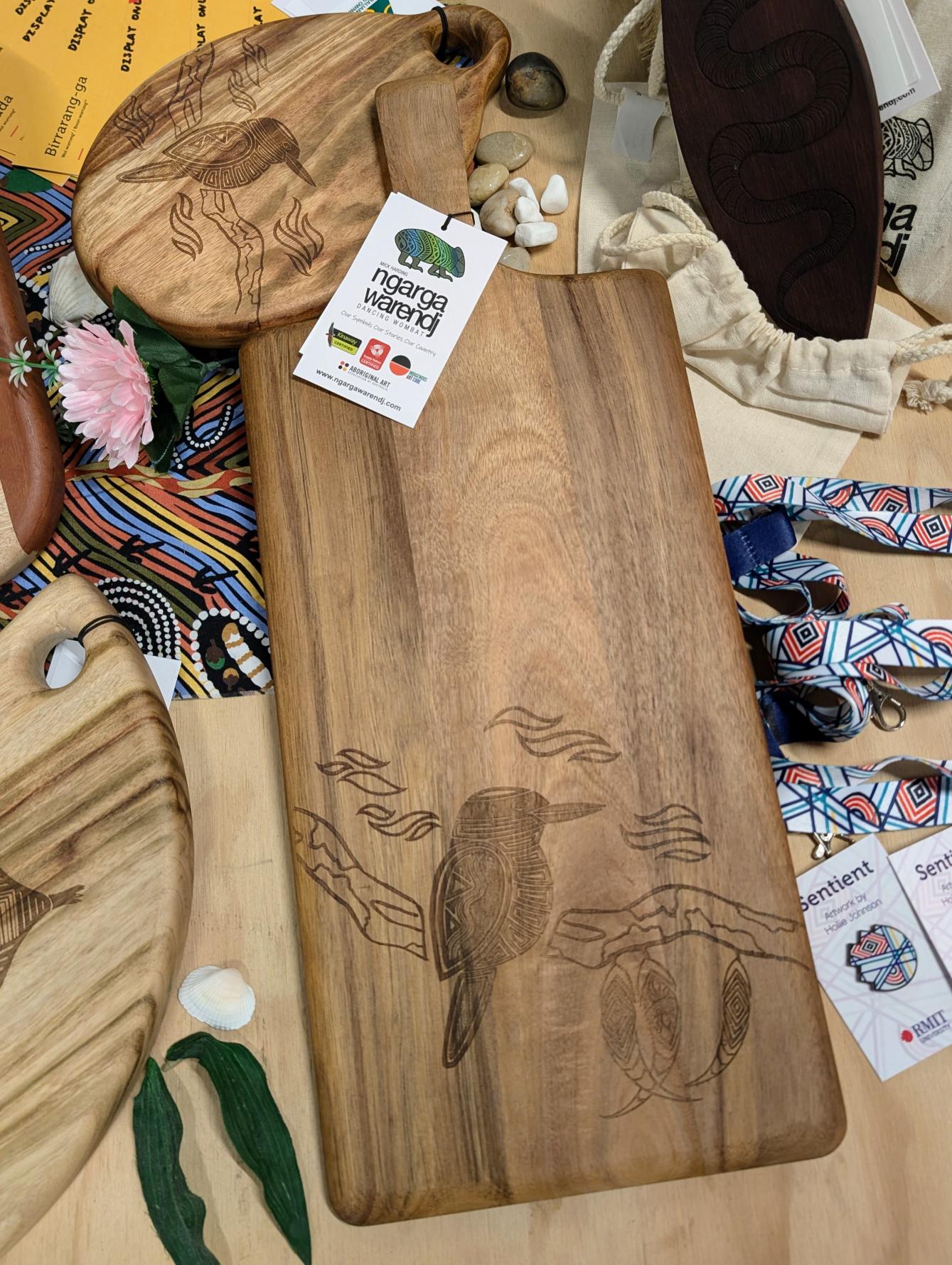 RMIT Store Acacia Rectangle Paddle Board, displayed on a wooden table in-store. The board features a smooth acacia wood surface with natural grain patterns and a rectangular shape with rounded edges. A detailed Kookaburra design is meticulously etched into the board, inspired by Traditional South Eastern Aboriginal art, adding a cultural touch to this functional and artistic piece. The piece is part of the Ngarga Warendj – Dancing Wombat collection.