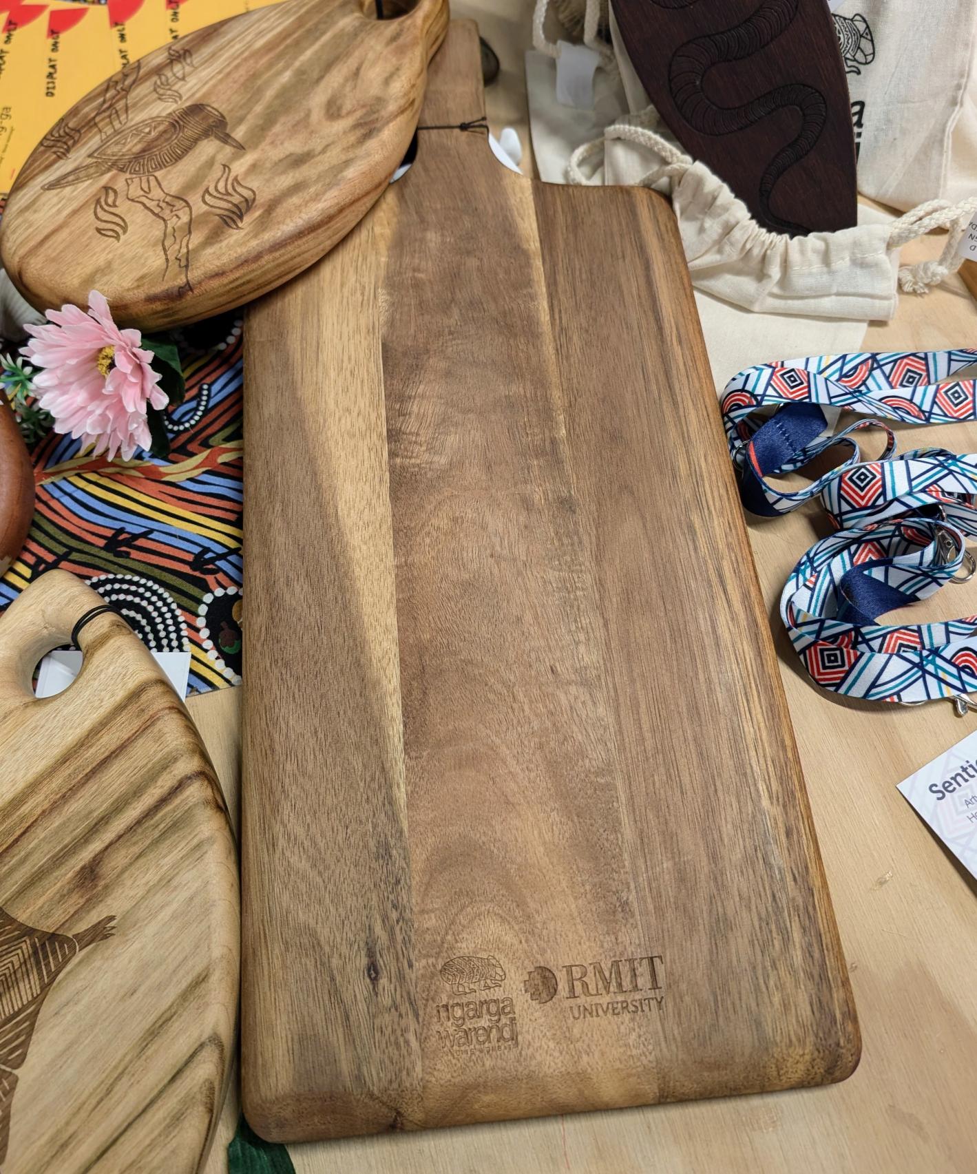 RMIT Store Acacia Rectangle Paddle Board, rear view displayed on a wooden table in-store. The acacia wood surface showcases the natural grain and smooth finish. Engraved at the bottom of the board is the RMIT University logo and the Ngarga Warendj branding, subtly marking the board’s cultural significance. This rear view highlights the board's simplistic, functional design, with a focus on the natural beauty of the acacia wood.