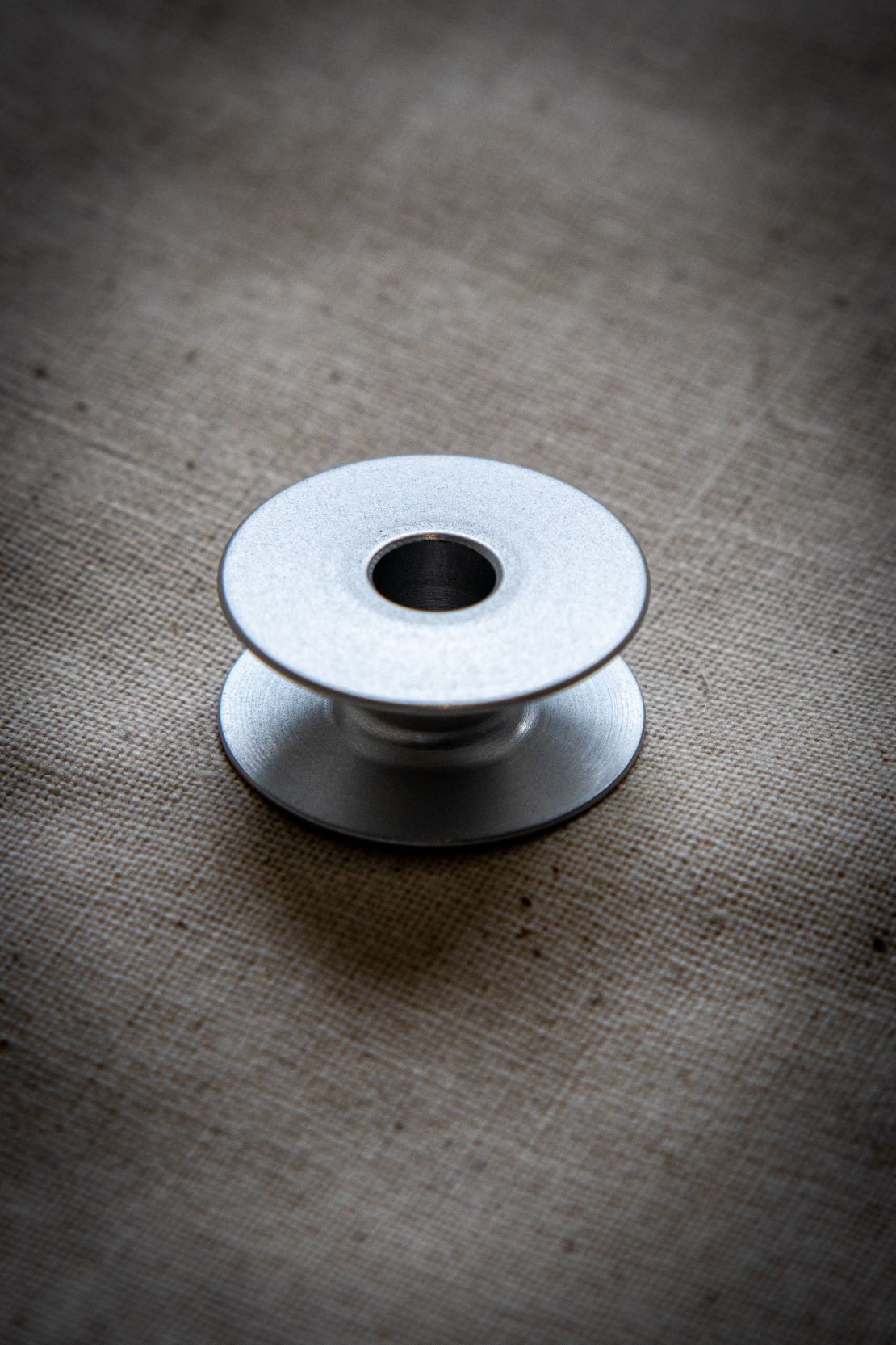 Top-down view of a single alloy bobbin lying flat on a light grey surface, clearly showing the symmetrical circular holes around the perimeter and a brushed metal texture. Available from the RMIT Store.