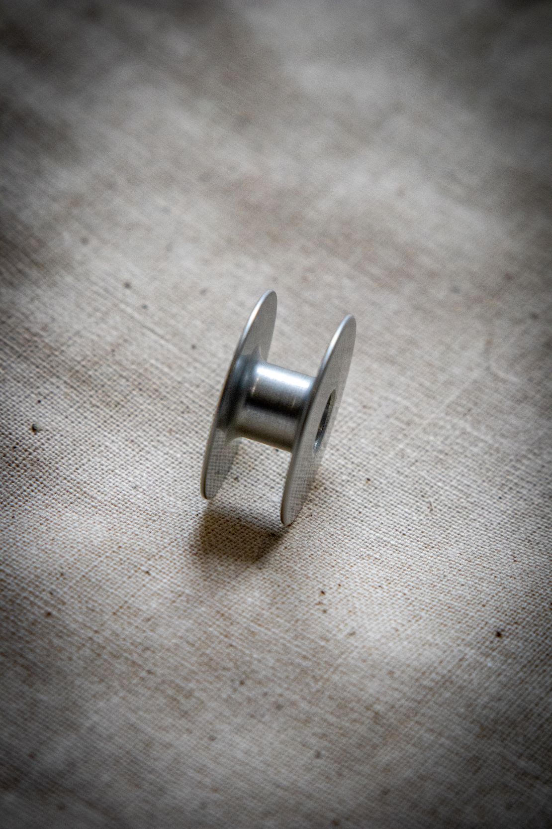 Side view of a single alloy bobbin standing upright on a light grey surface, featuring circular ventilation holes and a smooth metal finish.