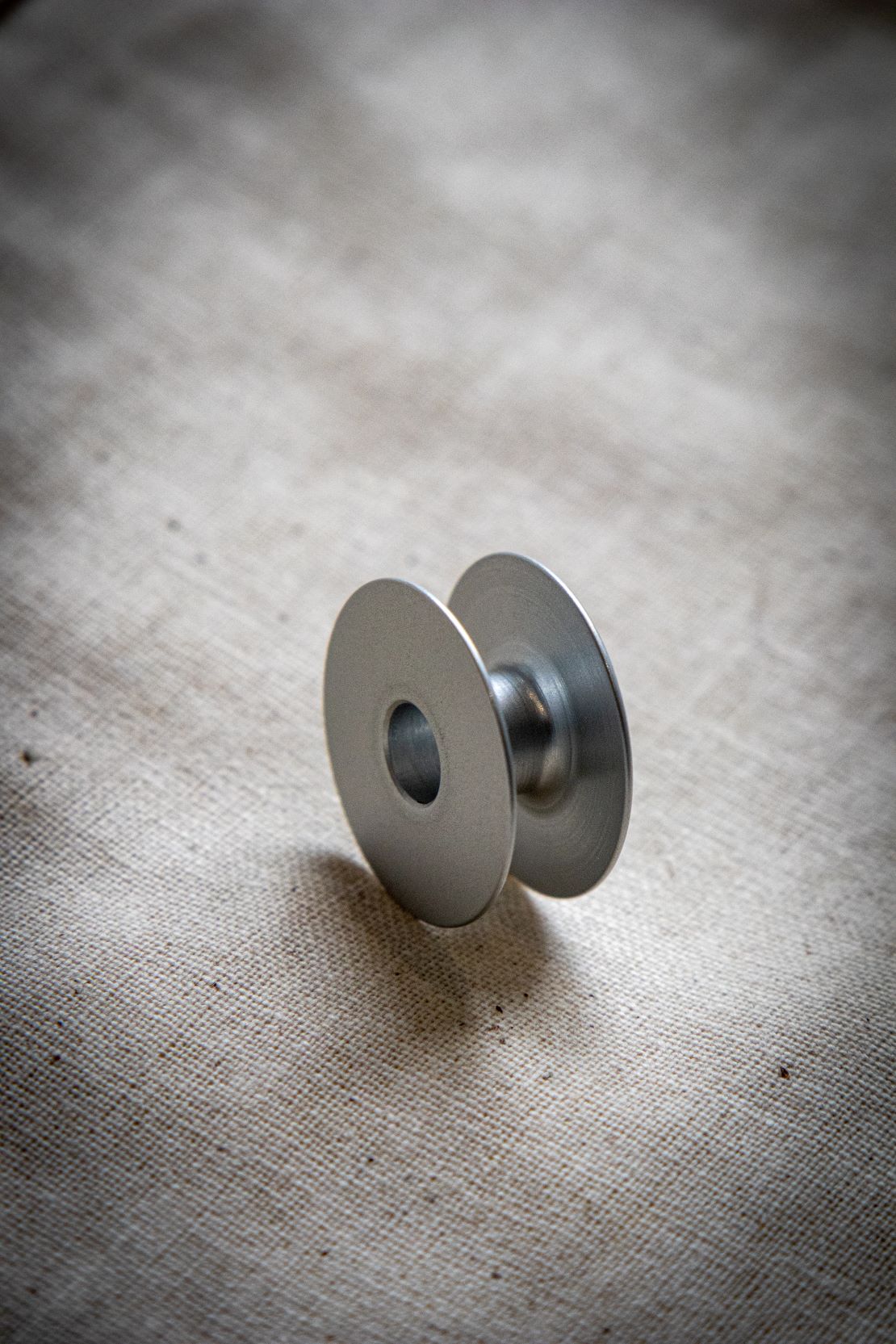 Close-up view of a single alloy bobbin shown upright on a light grey surface, displaying its smooth metal finish with circular side holes. Available from the RMIT Store.
