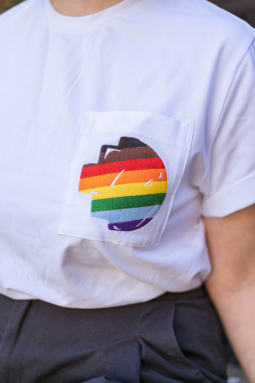 Close-up view of the left chest pocket on the RMIT Store Ally Tee in plain white, showing the pixel-style RMIT University logo with pride flag rainbow colours intricately embroidered on the fabric.