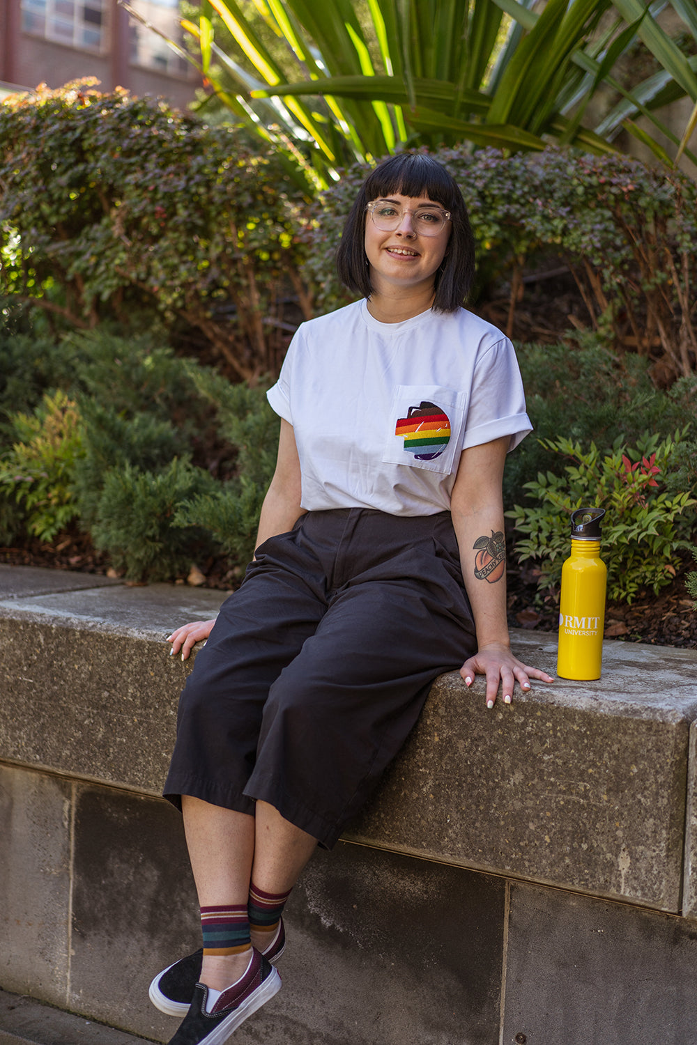 Close-up front view of a model seated on a stone bench wearing the white Ally Tee from the RMIT Store, showing the rainbow-coloured embroidered pixel logo on the chest pocket; a yellow water bottle is placed beside the model, with plants visible in the background.