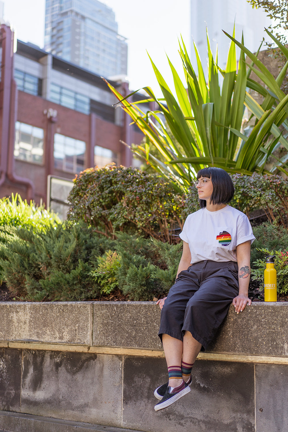 Model wearing the Ally Tee in white, showing the left chest pocket with the embroidered rainbow-coloured pixel logo, seated on a stone bench beside green plants in an outdoor setting, photographed for the RMIT Store.