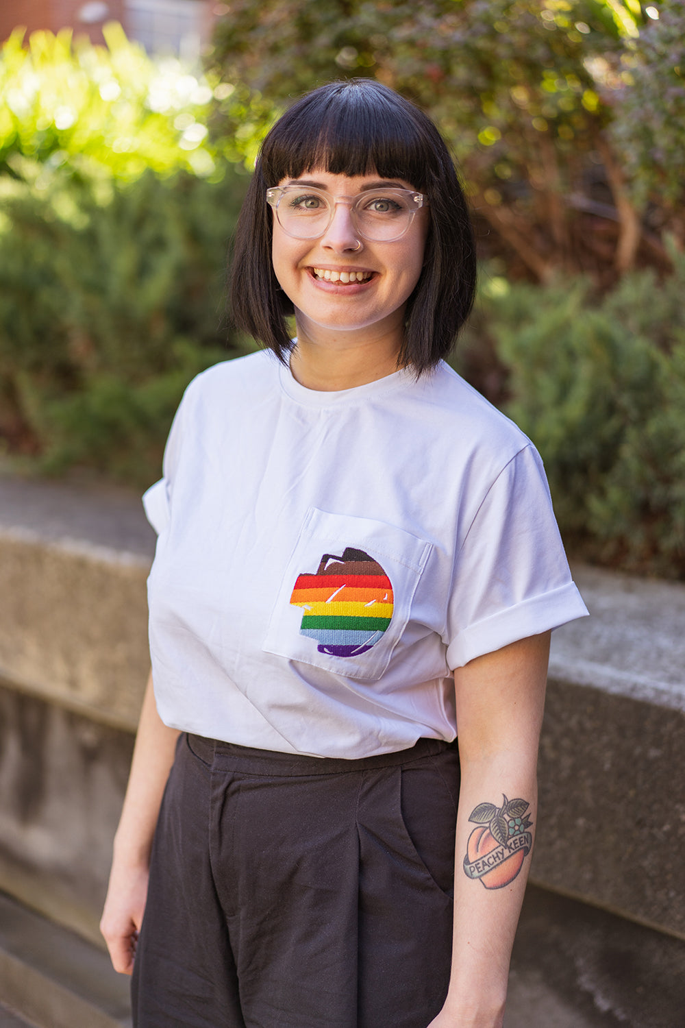Front view of a smiling model standing and wearing the white Ally Tee from the RMIT Store, showing the chest pocket with the rainbow-coloured embroidered pixel logo, against a background of greenery.