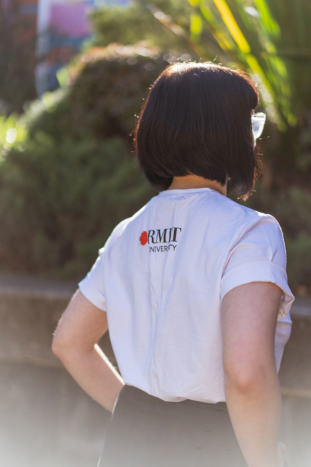 Side back view of a model wearing the white Ally Tee from the RMIT Store, featuring the plain white fabric with the RMIT University logo printed in black and red near the collar.