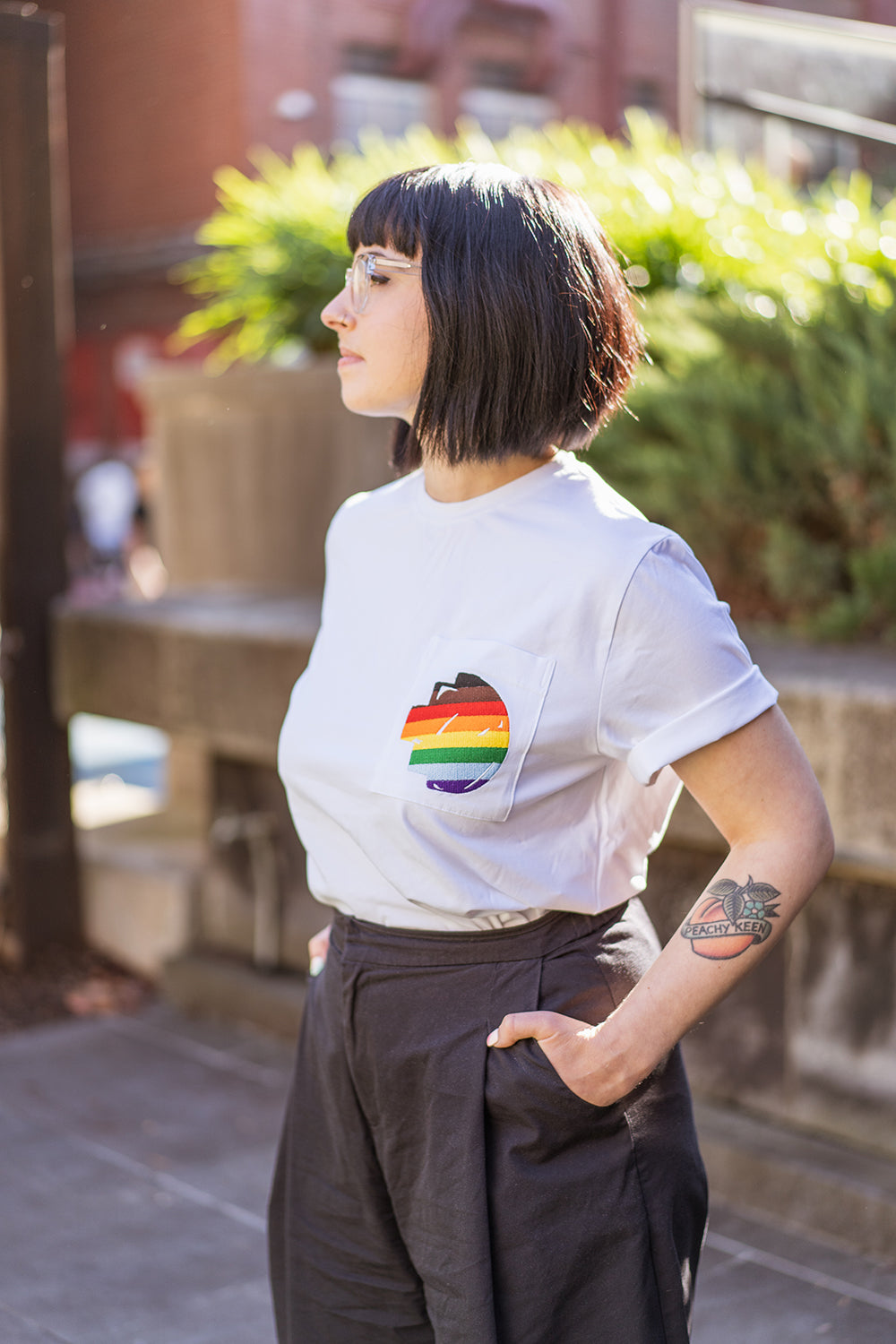 Side view of a model standing outdoors in sunlight wearing the white Ally Tee from the RMIT Store, with one hand in her pants pocket and the side of the T-shirt clearly visible, showing the chest pocket featuring a rainbow-coloured embroidered pixel logo; background includes natural greenery.