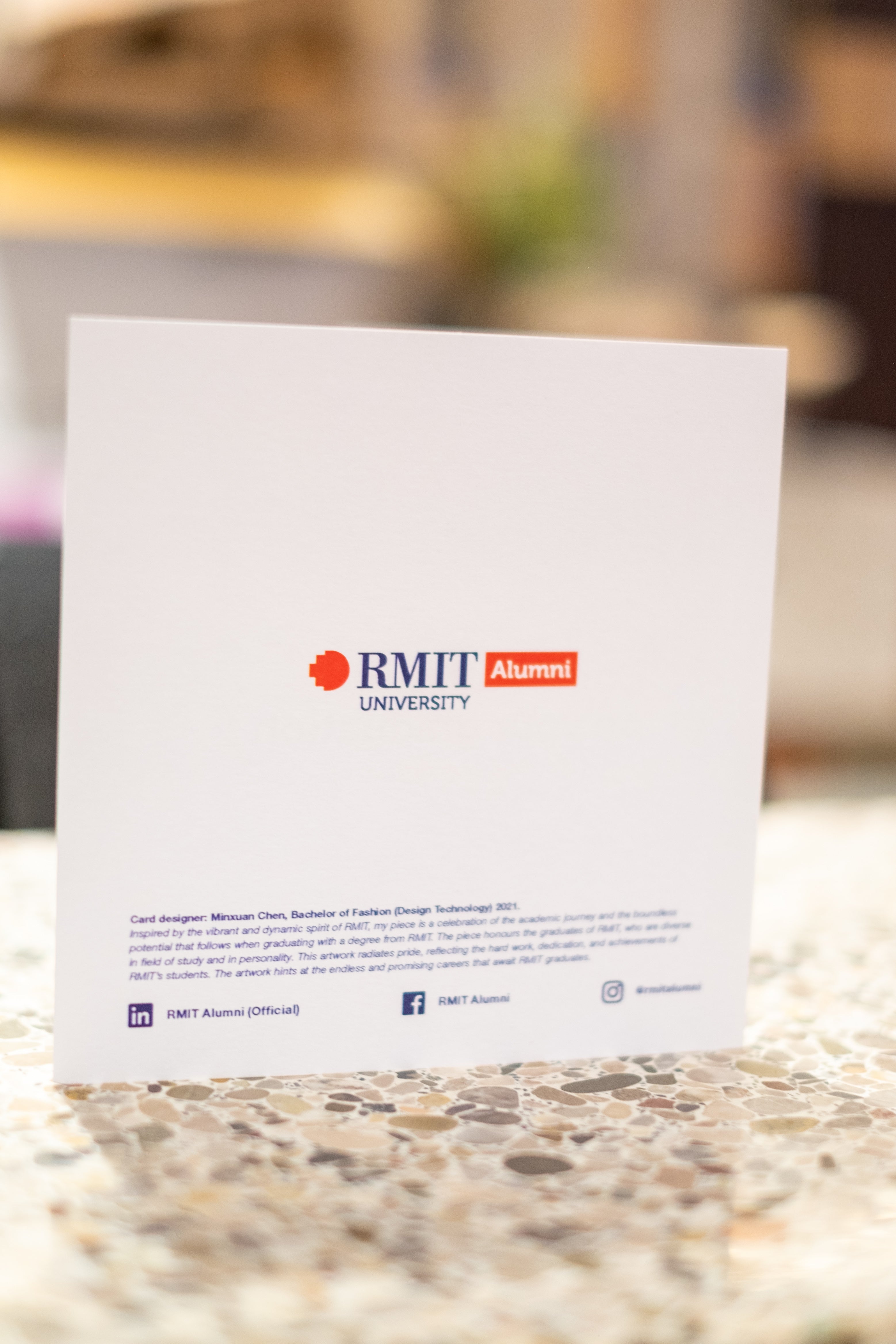 Back view of the alumni graduation card standing upright on a light tabletop, featuring the RMIT Alumni logo in navy and red centred on the card, available at the RMIT Store.