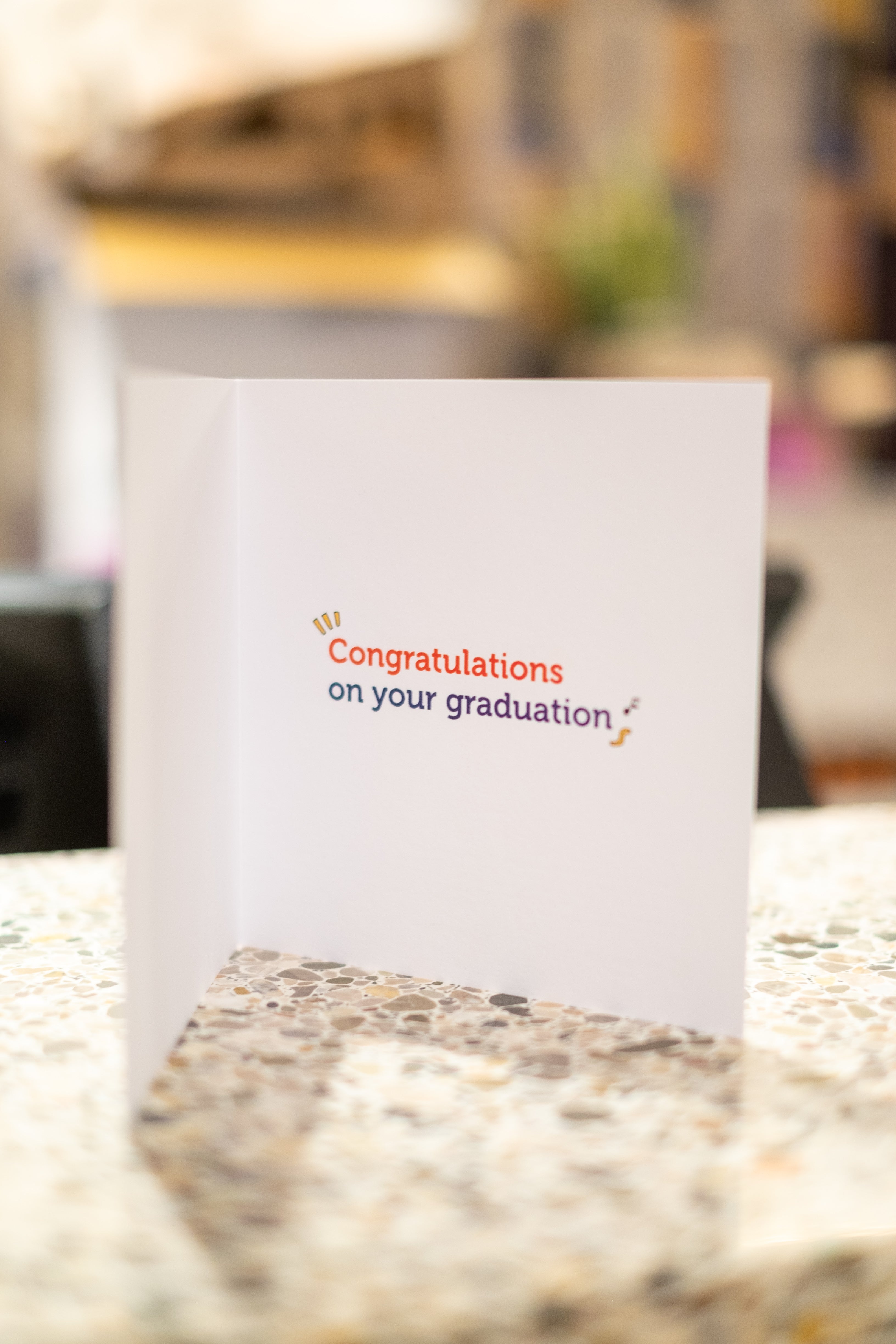 Inside view of the white RMIT Alumni graduation card ajar on a light tabletop. The card features the message 'Congratulations on your graduation' with 'Congratulations' in red and 'graduation' in navy, available at the RMIT Store.
